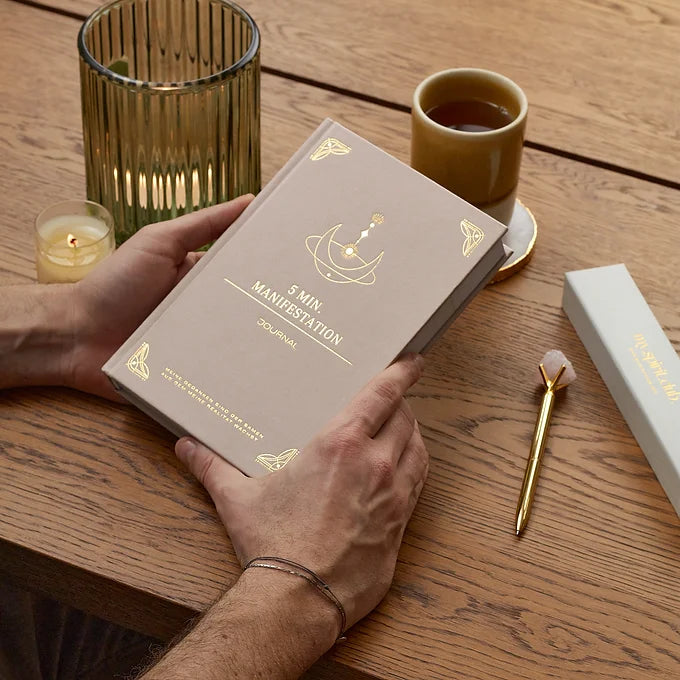 Man hält 5 Minuten Manifestation Journal in Beige auf einem Holztisch mit Kristall Stift